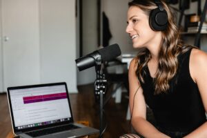 Podcaster wearing headphones and speaking into a microphone beside a laptop.