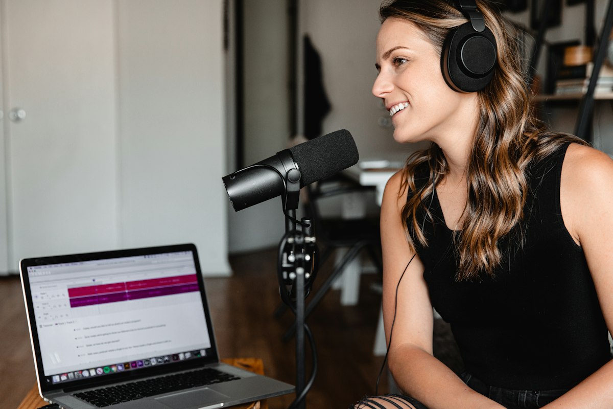 Podcaster wearing headphones and speaking into a microphone beside a laptop.